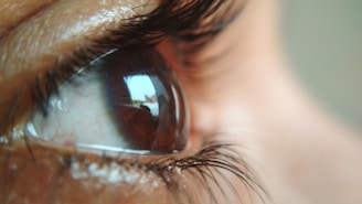 A close-up view of a human eye with detailed focus on the eyelashes and the reflection in the pupil. The reflections show a blurred, abstract pattern, suggesting a distorted view of an environment.