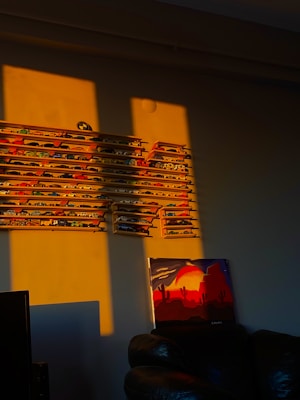 A wall display featuring numerous miniature car models, neatly arranged on several horizontal shelves. A colorful painting depicting a desert landscape with cacti and a setting sun is placed below the cars, resting against the wall. The room is illuminated by warm, golden sunlight casting long shadows on the wall and floor.