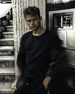 A man is leaning against a wall with a serious expression. He is wearing a black shirt and jacket. The background features a staircase and a metallic surface with some text in Spanish.