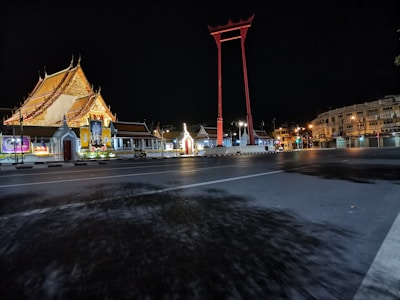 A large traditional Thai temple with an intricately designed golden roof is brightly illuminated at night. In the foreground, a main road is visible, leading to a red giant swing structure. The area is calm and devoid of people, with architectural buildings in the background further enhancing the scene.