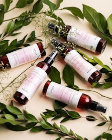 Five bottles of a skincare product with maroon-colored liquid and white labels are artistically arranged. They are surrounded by fresh green leaves and delicate white flowers, creating a natural and organic aesthetic.