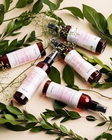 Five bottles of a skincare product with maroon-colored liquid and white labels are artistically arranged. They are surrounded by fresh green leaves and delicate white flowers, creating a natural and organic aesthetic.