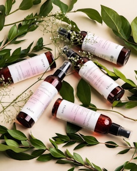 Five bottles of a skincare product with maroon-colored liquid and white labels are artistically arranged. They are surrounded by fresh green leaves and delicate white flowers, creating a natural and organic aesthetic.