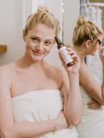 A person wrapped in a white towel is holding a bottle of skincare product next to their face, smiling gently. In the background, a reflection in the mirror shows the person applying makeup.