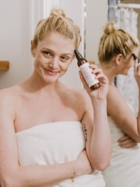 A person wrapped in a white towel is holding a bottle of skincare product next to their face, smiling gently. In the background, a reflection in the mirror shows the person applying makeup.