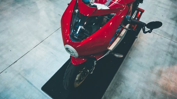 A red sports motorcycle is positioned on a display platform. The sleek design features sharp angles and glossy surfaces, highlighting its aerodynamic shape. The surrounding area appears to be an indoor showroom with a smooth, light-colored floor.