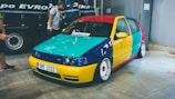 A small hatchback car with a custom paint job featuring bright, contrasting colors, including yellow, red, blue, and turquoise. It is parked indoors on a concrete floor with a reflective surface. The car has modified rims and low-profile tires. Two people are nearby, one of whom is pointing towards the car.