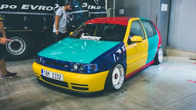 A small hatchback car with a custom paint job featuring bright, contrasting colors, including yellow, red, blue, and turquoise. It is parked indoors on a concrete floor with a reflective surface. The car has modified rims and low-profile tires. Two people are nearby, one of whom is pointing towards the car.