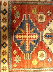 A traditional rug features intricate geometric patterns with a mix of vibrant and earthy tones. The dominant pattern includes a large central motif with diamond shapes and crosses, surrounded by smaller star and floral designs. The border has a repetitive series of these motifs, adding to the symmetry and complexity.