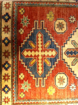 A traditional rug features intricate geometric patterns with a mix of vibrant and earthy tones. The dominant pattern includes a large central motif with diamond shapes and crosses, surrounded by smaller star and floral designs. The border has a repetitive series of these motifs, adding to the symmetry and complexity.