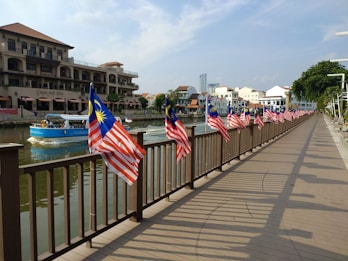 A wooden walkway lined with Malaysian flags stretches alongside a river. There are buildings with a mix of traditional and modern architectural styles on the opposite bank. A blue boat with passengers is navigating the waterway.
