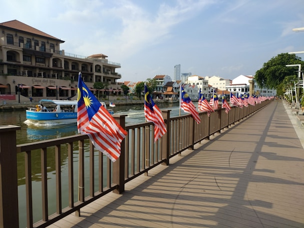 A wooden walkway lined with Malaysian flags stretches alongside a river. There are buildings with a mix of traditional and modern architectural styles on the opposite bank. A blue boat with passengers is navigating the waterway.