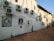 A row of air conditioning units is mounted on the exterior of a large, weathered white wall. The wall features several paintings, including portraits of individuals framed as if they are real windows or pictures. The ground is paved with orange-brown tiles, and the area appears to be an alley or side street with a clear blue sky overhead.
