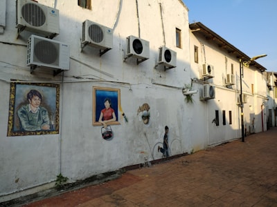 A row of air conditioning units is mounted on the exterior of a large, weathered white wall. The wall features several paintings, including portraits of individuals framed as if they are real windows or pictures. The ground is paved with orange-brown tiles, and the area appears to be an alley or side street with a clear blue sky overhead.