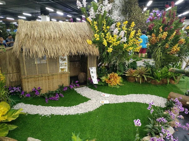 A rustic hut with a thatched roof is surrounded by a lush garden filled with vibrant tropical flowers and plants. The scene includes a path made of white pebbles meandering through the greenery, adding to the tranquil and natural setting. Colorful orchids and other floral arrangements contribute to the decorative display.