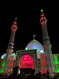 A majestic mosque brightly illuminated at night with intricate architectural designs. The mosaic tile patterns on two tall minarets and a large central dome are highlighted by red and green lights. A large crowd of people is gathered in front of the mosque, suggesting an event or celebration.