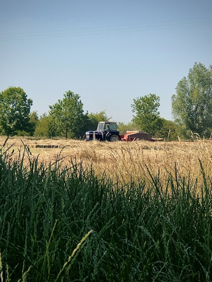 A rural landscape features a tractor in a field surrounded by trees. The foreground has tall, green grass, while the background includes the open field with hay or grain. Several trees are scattered, providing elements of greenery and shade.