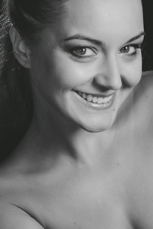 A close-up black and white portrait of a smiling person with light skin. The person is looking directly at the camera, conveying a sense of warmth and confidence. The image focuses on facial features, with particular emphasis on the eyes and smile.