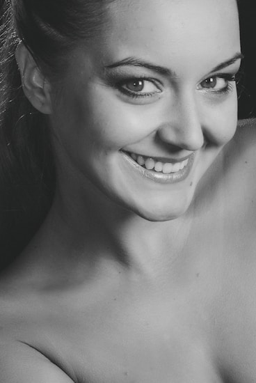 A close-up black and white portrait of a smiling person with light skin. The person is looking directly at the camera, conveying a sense of warmth and confidence. The image focuses on facial features, with particular emphasis on the eyes and smile.