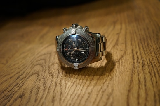 A silver luxury wristwatch with a dark, detailed dial featuring multiple sub-dials and a metal bracelet is placed on a wooden surface. The watch has a polished bezel and luminous hour markers, and the watch hands are prominently visible.