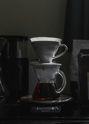 A pour-over coffee setup with a white ceramic dripper placed on top of a glass carafe. The carafe contains freshly brewed coffee and rests on a digital kitchen scale. The scene is dimly lit, creating a cozy and focused atmosphere.