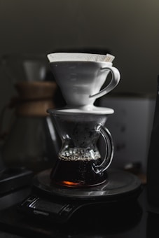 A pour-over coffee maker with a white ceramic dripper and a transparent carafe is filled with freshly brewed coffee, placed on a digital kitchen scale. The scene is dimly lit, creating a warm and cozy atmosphere.