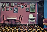 A retro-themed room featuring a wall adorned with framed photographs and vintage records. A small dining table with red cushioned chairs sits against the wall. The floor is covered with a pattern resembling vinyl records, and a high chair is positioned nearby. The room has a nostalgic and eclectic ambiance, with a mix of pink and blue walls.
