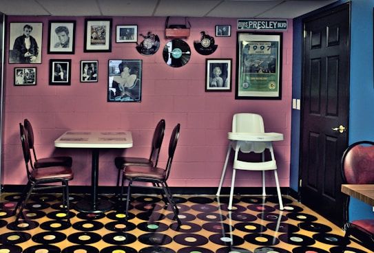 A retro-themed room featuring a wall adorned with framed photographs and vintage records. A small dining table with red cushioned chairs sits against the wall. The floor is covered with a pattern resembling vinyl records, and a high chair is positioned nearby. The room has a nostalgic and eclectic ambiance, with a mix of pink and blue walls.