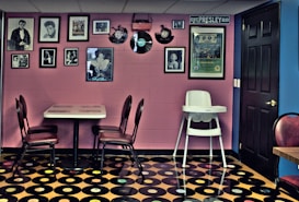 A retro-themed room featuring a wall adorned with framed photographs and vintage records. A small dining table with red cushioned chairs sits against the wall. The floor is covered with a pattern resembling vinyl records, and a high chair is positioned nearby. The room has a nostalgic and eclectic ambiance, with a mix of pink and blue walls.