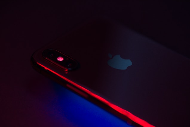 A close-up of a smartphone with a prominent Apple logo, illuminated by red and blue lighting. The camera lens and edges are highlighted, creating a futuristic and sleek aesthetic.