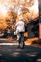 An older woman wearing a floral patterned outfit rides a bicycle down a quiet, tree-lined street. The trees display vibrant autumn foliage, casting warm golden hues over the scene. A building with a brick exterior is visible on the right, and a traffic sign is seen along the roadway.