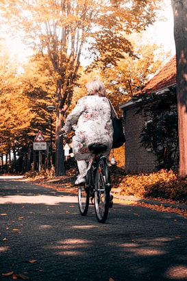 An older woman wearing a floral patterned outfit rides a bicycle down a quiet, tree-lined street. The trees display vibrant autumn foliage, casting warm golden hues over the scene. A building with a brick exterior is visible on the right, and a traffic sign is seen along the roadway.