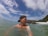 A person wearing a backward cap is in the water near a rocky coastline. In the background, a speedboat is floating on the water, and there is a small island with vegetation visible. The sky is partly cloudy, and the water appears calm.