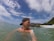 A person wearing a backward cap is in the water near a rocky coastline. In the background, a speedboat is floating on the water, and there is a small island with vegetation visible. The sky is partly cloudy, and the water appears calm.
