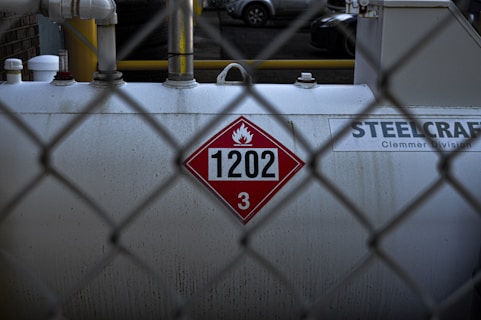 A large industrial tank is positioned behind a chain-link fence. The tank displays a hazardous materials placard with the number 1202 and a flammable symbol. The surface appears worn and slightly stained. Pipes and fittings are visible above the tank, and the background shows parts of parked vehicles.