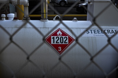 A large industrial tank is positioned behind a chain-link fence. The tank displays a hazardous materials placard with the number 1202 and a flammable symbol. The surface appears worn and slightly stained. Pipes and fittings are visible above the tank, and the background shows parts of parked vehicles.