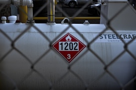 A large industrial tank is positioned behind a chain-link fence. The tank displays a hazardous materials placard with the number 1202 and a flammable symbol. The surface appears worn and slightly stained. Pipes and fittings are visible above the tank, and the background shows parts of parked vehicles.