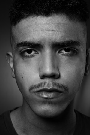 A close-up black and white portrait of a young man with a serious expression. The image highlights his facial features including a mustache and short hair.