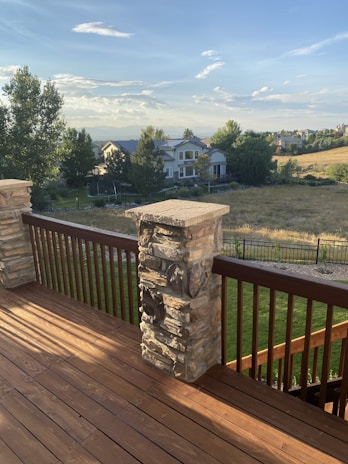 A wooden deck with stone pillars is overlooking a grassy field and residential houses in the distance. The sky is partly cloudy, with the setting sun casting warm tones on the landscape.