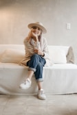 A person is seated on a white couch in a minimalistic room. They wear a light-colored hat, a beige sweater, rolled-up jeans, and white sneakers. The setting conveys a relaxed and cozy atmosphere, with soft lighting and neutral-toned walls.