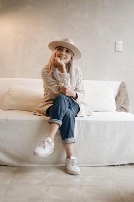 A person is seated on a white couch in a minimalistic room. They wear a light-colored hat, a beige sweater, rolled-up jeans, and white sneakers. The setting conveys a relaxed and cozy atmosphere, with soft lighting and neutral-toned walls.