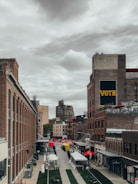 A vibrant urban scene with rows of brick buildings lining a cobblestone street, leading into the distance. Red and yellow market tents are set up along the street, suggesting an outdoor market or fair. A prominent sign on the building reads 'VOTE' in large letters. The sky is overcast, casting a muted light over the scene.