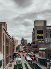A vibrant urban scene with rows of brick buildings lining a cobblestone street, leading into the distance. Red and yellow market tents are set up along the street, suggesting an outdoor market or fair. A prominent sign on the building reads 'VOTE' in large letters. The sky is overcast, casting a muted light over the scene.