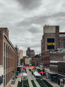 A vibrant urban scene with rows of brick buildings lining a cobblestone street, leading into the distance. Red and yellow market tents are set up along the street, suggesting an outdoor market or fair. A prominent sign on the building reads 'VOTE' in large letters. The sky is overcast, casting a muted light over the scene.