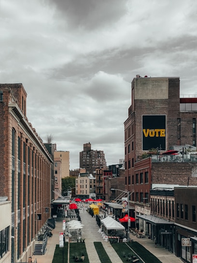 A vibrant urban scene with rows of brick buildings lining a cobblestone street, leading into the distance. Red and yellow market tents are set up along the street, suggesting an outdoor market or fair. A prominent sign on the building reads 'VOTE' in large letters. The sky is overcast, casting a muted light over the scene.
