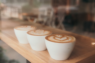 Three white cups of coffee with intricate latte art sit on a wooden surface. The design on the foam features heart and leaf patterns. The background is blurred, suggesting a cozy café setting.