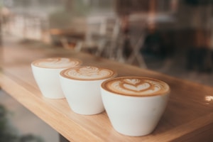 Three white cups of coffee with intricate latte art sit on a wooden surface. The design on the foam features heart and leaf patterns. The background is blurred, suggesting a cozy café setting.