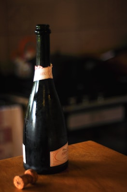 A dark glass bottle of sparkling wine with a beige and pink label stands on a wooden table. Beside the bottle is a cork, indicating that the bottle has been opened. The background appears to be a dimly lit kitchen or bar setting, with blurred outlines of kitchen appliances.