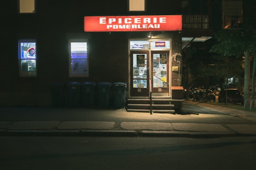 A small convenience store with a bright red sign displaying the name 'Épicerie Pomerleau' above the entrance. The storefront is illuminated, contrasting with the dimly lit surroundings. There are display windows featuring various advertisements and two doors with multiple flyers attached. A row of recycling bins is positioned to the left of the entrance.