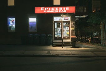 A small convenience store with a bright red sign displaying the name 'Épicerie Pomerleau' above the entrance. The storefront is illuminated, contrasting with the dimly lit surroundings. There are display windows featuring various advertisements and two doors with multiple flyers attached. A row of recycling bins is positioned to the left of the entrance.
